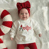 Baby wearing a white sweatshirt with 'Amilee' and red pants, lying on a textured surface next to a candy cane.