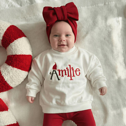 Baby wearing a white sweatshirt with 'Amilee' and red pants, lying on a textured surface next to a candy cane.