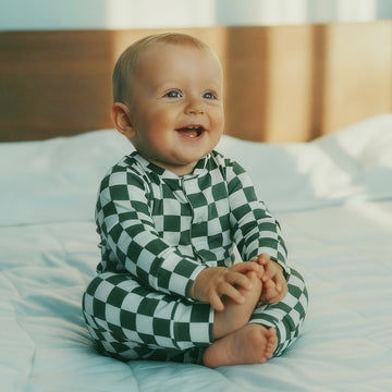 Baby wearing a green and white checkered outfit sitting on a bed.