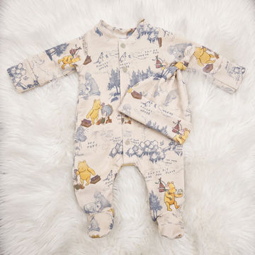 A baby footed sleeper and matching hat laid flat on a white faux fur background. The sleeper is cream-colored with a vintage-style travel map print featuring Winnie the Pooh characters and blue-gray illustrations, with a matching hat 