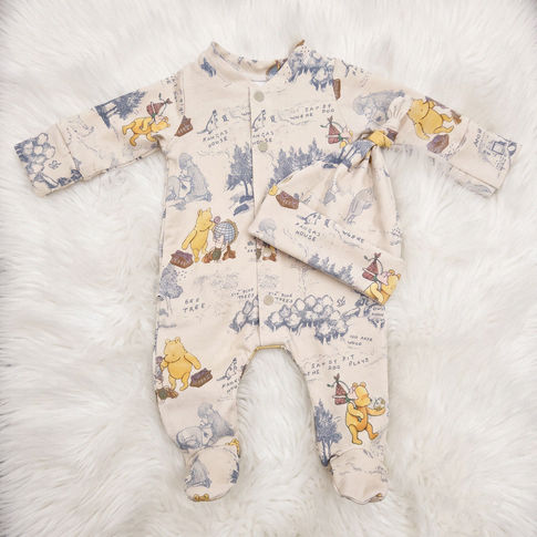 A baby footed sleeper and matching hat laid flat on a white faux fur background. The sleeper is cream-colored with a vintage-style travel map print featuring Winnie the Pooh characters and blue-gray illustrations, with a matching hat 