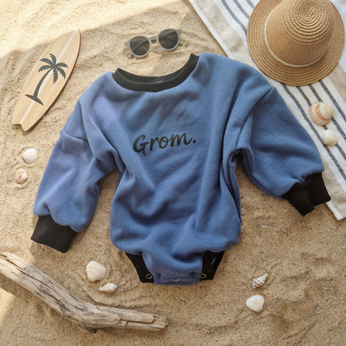 Blue sweatshirt with 'Grom' text on a sandy surface with beach accessories.