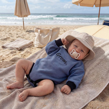 Baby on a beach with a pacifier, wearing a blue outfit and beige hat.