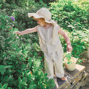 Child in a garden wearing a sun hat and striped outfit, holding a basket.
