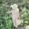 Little girl in a sun hat and cotton top and trouser set standing among green foliage