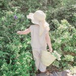 Little girl in a sun hat and cotton top and trouser set standing among green foliage