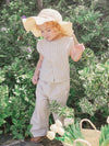 Child in a beige outfit with a wide-brimmed hat standing among green foliage.