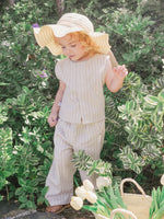 Child in a beige outfit with a wide-brimmed hat standing among green foliage.