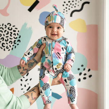 Baby in a colorful outfit with abstract patterns, held by a person against a patterned background.