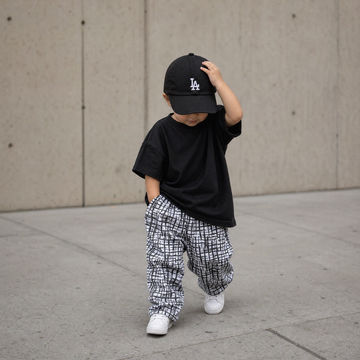 Child wearing a black cap and black shirt with white pants, standing on a concrete surface.