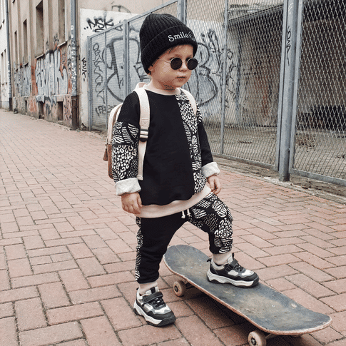 Toddler wearing black and white leopard print tracksuit, with smile beanie and black sunglasses. Standing on a skateboard in a street, urban scene. 