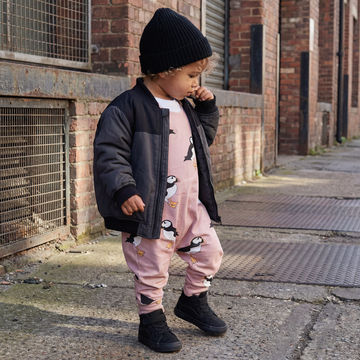 Toddler boy wearing taupe puffins dungarees with an oversized black jacket and beanie hat.