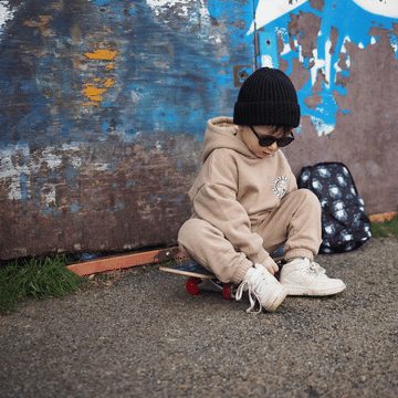 toddler boy wearing beige hooded tracksuit with black accessories. Sitting on a skateboard in an urban street scene.
