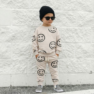 Child wearing a beige outfit with smiley face patterns against a white brick wall.