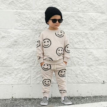 Child wearing a beige outfit with smiley face patterns against a white brick wall.