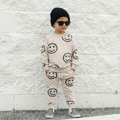 Child wearing a beige outfit with smiley face patterns against a white brick wall.