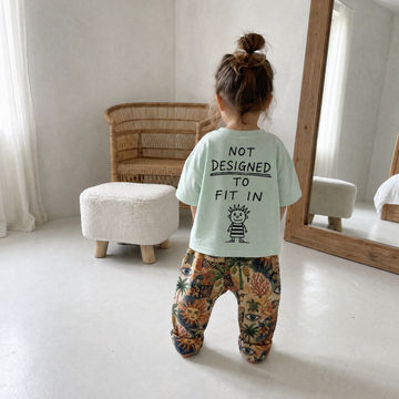 A toddler stands with their back to the camera in a bright, minimalist room. The child is wearing a light mint green t-shirt with black text that reads 'NOT DESIGNED TO FIT IN' and a small cartoon character, paired with colorful, patterned pants featuring a tropical floral and palm tree design.