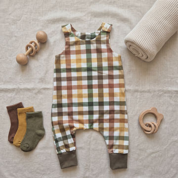 A baby romper with a warm-toned plaid pattern in mustard yellow, brown, green, and white, styled in a flat lay on a beige linen background with baby accessories including wooden teething rings, socks, and a rolled blanket.