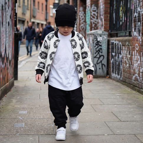 Child walking on a street with graffiti-covered walls wearing lion print Bomber jacket, black trousers and beanie hat. 