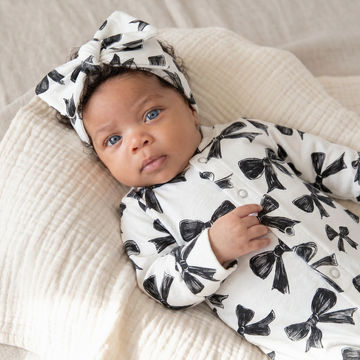 A baby lying on a textured cream muslin blanket, wearing a white long-sleeve bodysuit and matching bow-patterned headband.