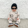 Baby wearing a beige outfit with smiley face patterns, black sunglasses, and a black beanie against a white brick wall.