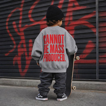 A child standing with a skateboard in front of a black metal shutter with red graffiti, wearing a gray sweatshirt with bold red text that reads 'CANNOT BE MASS PRODUCED.'.