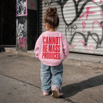 A toddler viewed from behind wearing a light pink sweatshirt with bold red text that reads 'CANNOT BE MASS PRODUCED.' and light blue jeans, walking outdoors in front of a graffiti-covered metal shutter.