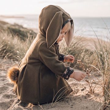 A toddler wearing a brown bunny costume with long floppy ears, a fluffy tail, and patterned cuffs, crouching on a sandy beach dune with grass and the ocean in the background.