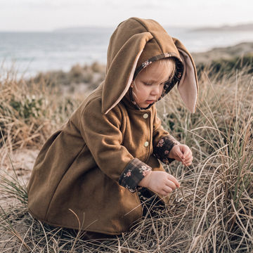 A toddler wearing a brown bunny hooded coat sits in dry beach grass with the ocean blurred in the background.