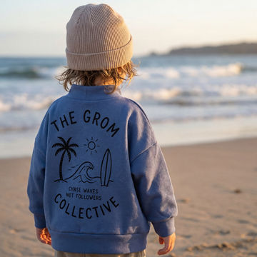 Child wearing a blue jacket with 'The Grom Collective' design on a beach.