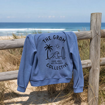 Blue sweatshirt with 'The Grom Collective' design on a wooden fence at the beach.