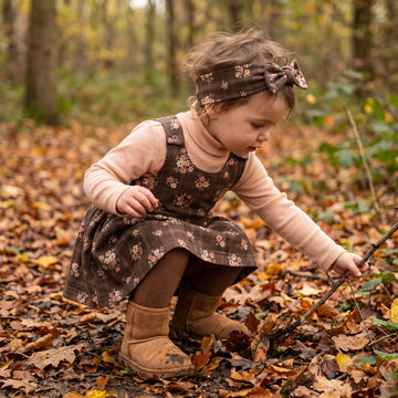 A toddler girl crouching in a wooded outdoor setting covered with fallen leaves, wearing a brown floral pinafore dress over a light beige turtleneck, brown tights, tan boots, and a matching headband.
