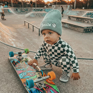 Child in checkered outfit and beanie with smiley face design at a skate park.