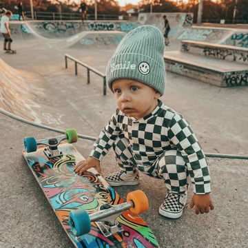 Child in checkered outfit and beanie with smiley face design at a skate park.