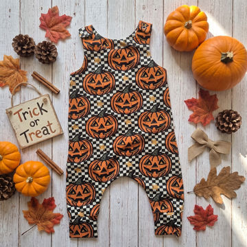 A baby romper with an all-over Halloween pumpkin print, styled in a flat lay on a white wood background with autumn props including pumpkins, fall leaves, pinecones, cinnamon sticks, and a 'Trick or Treat' sign.