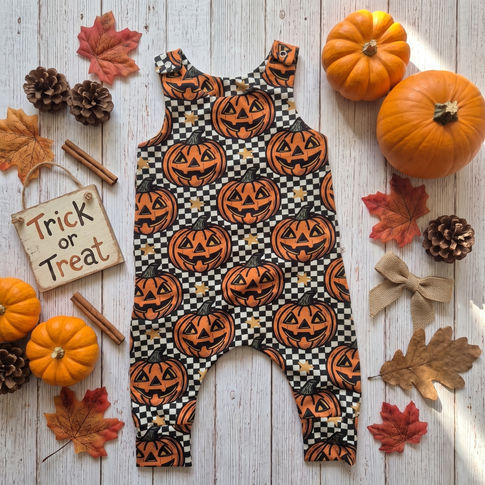 A baby romper with an all-over Halloween pumpkin print, styled in a flat lay on a white wood background with autumn props including pumpkins, fall leaves, pinecones, cinnamon sticks, and a 'Trick or Treat' sign.