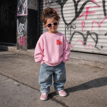 A young child wearing a pink crewneck sweatshirt with 'NAME YOUR DESIRE IN PRINCE' in red text, paired with baggy blue jeans and pink sneakers, standing outdoors in front of a graffiti-covered metal shutter.