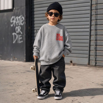 A child wearing a light gray sweatshirt with red text, black beanie, sunglasses, and black sneakers, holding a skateboard in front of a gray metal wall with graffiti.