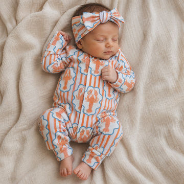 A sleeping newborn baby wearing a long-sleeve one-piece jumpsuit and matching headband, both in a light blue fabric with orange lobster illustrations and vertical orange stripes, laid on a beige textured blanket.