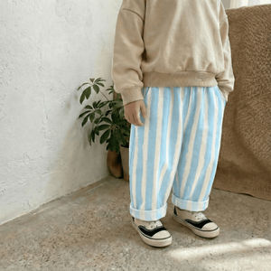 Person wearing a beige sweatshirt and blue and white striped pants standing against a white wall with a plant and brown cushion in the background.