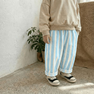 Person wearing a beige sweatshirt and blue and white striped pants standing against a white wall with a plant and brown cushion in the background.