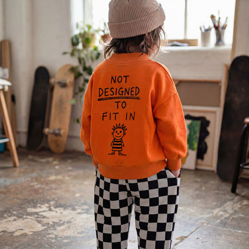 Child wearing an orange sweatshirt with a message and checkered pants in a room with skateboards.