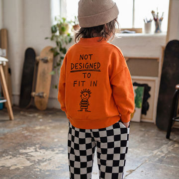 Child wearing an orange sweatshirt with a quote and graphic, standing in a room with skateboards and plants.