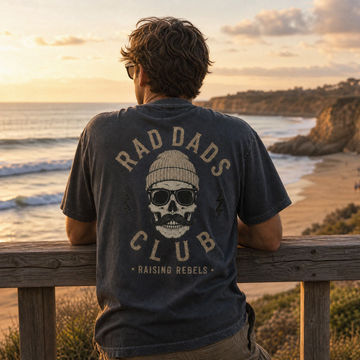 Man wearing a 'RAD DADS CLUB' t-shirt with a skull design, sitting on a wooden railing overlooking the ocean at sunset.