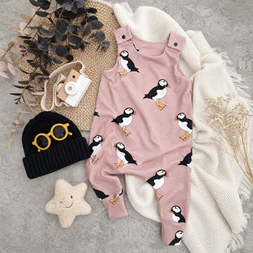 A baby romper in dusty pink with a repeating puffin print, styled in a flat lay with a cream knit blanket, a black beanie with yellow glasses, a cream star toy, and dried botanicals on a gray concrete surface.