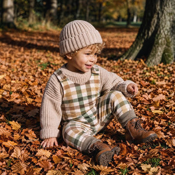 A toddler sitting on a forest floor covered in fallen autumn leaves, wearing a beige knit beanie, a cream and olive green plaid overall-style romper, and brown lace-up boots.