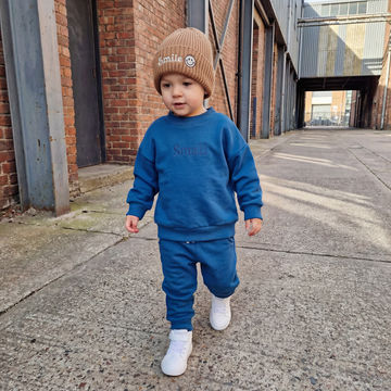 Child wearing a blue outfit and brown beanie with 'Smile' text, standing on a concrete surface.
