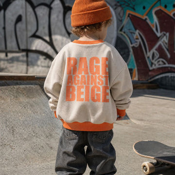 A child seen from behind wearing a cream sweatshirt with bold orange text reading 'RAGE AGAINST BEIGE' on the back, paired with dark jeans and an orange beanie, standing at a skatepark with a graffiti wall in the background.