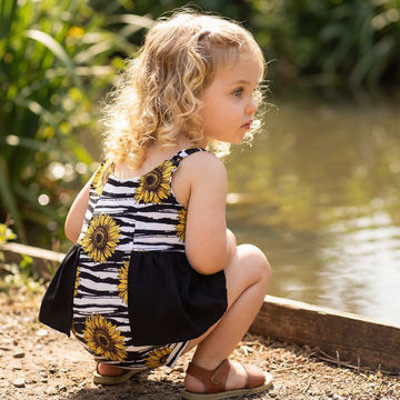 toddler girl wearing sunflowers print summer romper crouched down beside a lake.
