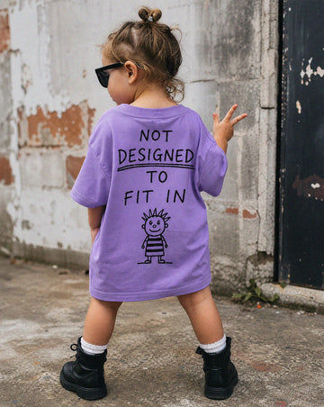 Child wearing a purple shirt with Not designed to fit in text and a graphic, standing against a textured wall.
